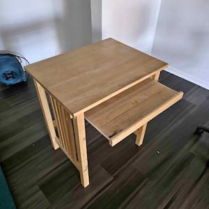 May include: A light-colored wooden side table with a pull-out tray. The table has a rectangular top and a lower shelf. The legs are sturdy and the design is simple and functional. The table is approximately 24 inches tall.