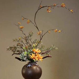 Puede incluir: Un arreglo floral en un jarrón de cerámica redondo de color marrón oscuro. La composición presenta lirios de color naranja brillante, racimos de pequeñas bayas de color verde claro y ramas finas y arqueadas con flores amarillas.