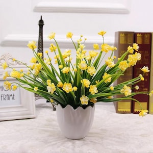 Op de afbeelding: Een boeket kleine gele bloemen met groene stengels en bladeren in een witte keramische pot met een geschulpte rand. De bloemen hebben meerdere bloemblaadjes en staan dicht bij elkaar in een ronde vorm met heldergele bloesems.