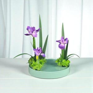 Könnte beinhalten: Ein rundes, hellgrünes Keramiktablett enthält ein Blumenarrangement mit drei violetten Schwertlilien mit gelben Akzenten, langen grünen Blättern und kleinen Büscheln aus hellgrünem Moos an der Basis jeder Blumengruppe.