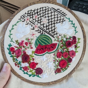 May include: Embroidered hoop art featuring a watermelon, red roses, white doves, and a black geometric design. The artwork is set against a cream-colored fabric, with a wooden hoop frame. The design includes green leaves and red accents.