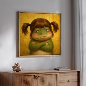Puede incluir: Pintura enmarcada de un personaje gruñón con forma de rana con los brazos cruzados. La rana tiene piel verde, mejillas rosadas y cabello castaño en coletas. El fondo es amarillo texturizado y el marco es dorado.
