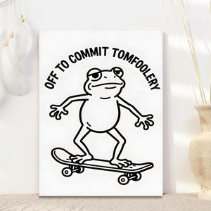 May include: A white print with a black outline illustration of a frog on a skateboard. The text "OFF TO COMMIT TOMFOOLERY" is above the frog. The image is on a white background.