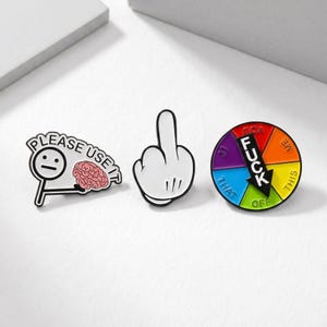 May include: Three enamel pins with black outlines. One pin features a stick figure with the text "PLEASE USE IT" and a brain graphic. Another pin is a white hand giving the middle finger. The third pin is a colourful wheel with the word "FUCK" and arrows.