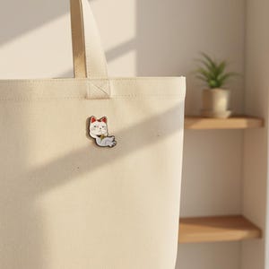 Op de afbeelding: Een wit en rode Maneki-neko kat emaille pin, met een gouden kraag, is bevestigd aan een beige draagtas. De pin toont een ontspannen kat, een symbool van geluk en voorspoed.