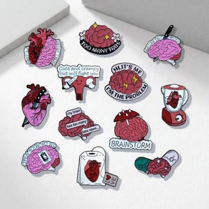 May include: A collection of enamel pins with various designs, including anatomical hearts, brains, and a uterus. Some pins feature text like "TOO MANY TABS," "BRAINSTORM," and "I'M THE PROBLEM." The pins are in shades of red, pink, and white.