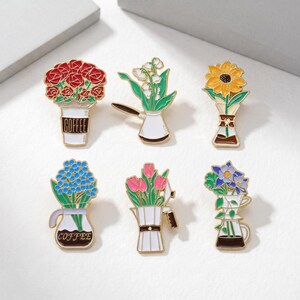 May include: A collection of six floral enamel pins. The pins feature various flower arrangements in coffee cups, coffee pots, and vases. Colors include red, yellow, blue, and pink, with gold and white accents. The coffee cup pin has the word "COFFEE" on it.