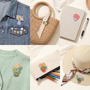 Op de afbeelding: Een verzameling kleurrijke emaille pins met bloementhema. De pins tonen verschillende bloemstukken in potten en vazen, bevestigd aan een spijkerjasje, een strooien tas, een notitieboek, een trui, een etui en een zonnehoed.