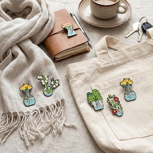 Op de afbeelding: Een assortiment kleurrijke emaille pins met bloemen- en plantontwerpen. De pins hebben de vorm van vazen met bloemen en planten. De pins liggen op een sjaal, een totebag en een leren notitieboek. Een kopje koffie, sleutels en een pen zijn ook zichtbaar.