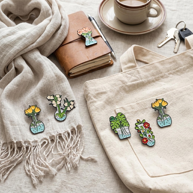 Op de afbeelding: Een assortiment kleurrijke emaille pins met bloemen- en plantontwerpen. De pins hebben de vorm van vazen met bloemen en planten. De pins liggen op een sjaal, een totebag en een leren notitieboek. Een kopje koffie, sleutels en een pen zijn ook zichtbaar.
