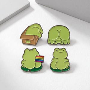 May include: Four green frog enamel pins. One frog is in a brown box, another is upside down, and a third holds a rainbow flag. The fourth frog is sitting on a lily pad. Each pin has a black outline.