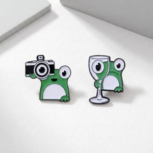 Może przedstawiać: Dwie emaliowane przypinki przedstawiające kreskówkowe żaby. Jedna żaba trzyma czarny aparat fotograficzny, druga kieliszek do wina. Obie żaby są zielono-białe z czarnymi akcentami. Przypinki są na białym tle.