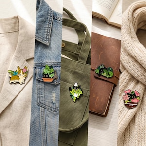 Op de afbeelding: Een verzameling emaille pins met diverse ontwerpen. Bevat een corgi met vleugels en een bloem, een potplant, een zwarte kat in een plant, een kat die in bladeren slaapt en een kat in een bloempot. De pins zijn bevestigd aan kleding en accessoires.