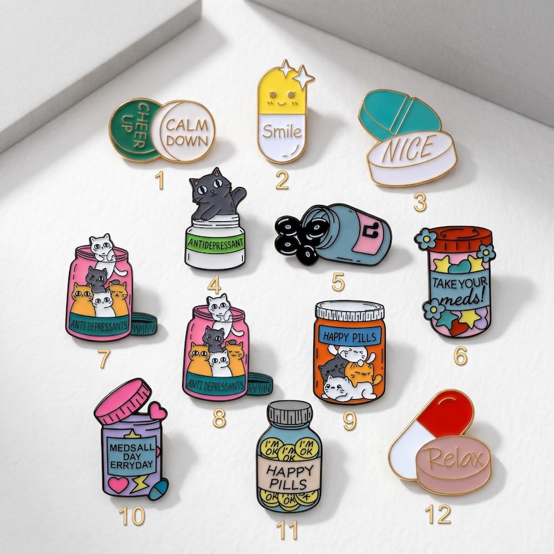 May include: A collection of colorful enamel pins with various designs. The pins include pills, cat illustrations, and text such as "Smile", "Nice", "Happy Pills", and "Relax". The pins are in various shapes and sizes.