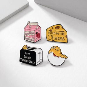 May include: Four enamel pins: a pink milk carton with a strawberry, a cheese wedge with text, a black and white toaster with text, and a yellow egg character. The milk carton says "MOOOOD" and "FRESH MILK". The cheese says "FOLD IN THE Cheese".
