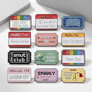 May include: A collection of enamel pins with various designs and text. The pins feature rectangular and ticket shapes with phrases like "Hello, I'm Super Gay" and "Romance Book Club." Colors include red, pink, green, and black.