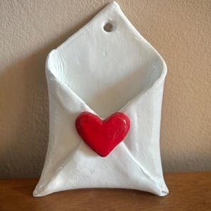 May include: A white ceramic wall pocket shaped like a folded envelope with a smooth, glossy finish. A small, three-dimensional red heart is attached to the center of the envelope flap. A circular hole is positioned at the top point for hanging.