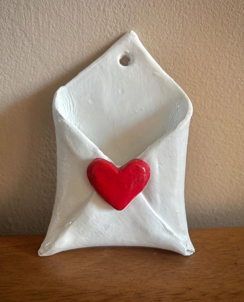 May include: A white ceramic wall pocket shaped like a folded envelope with a smooth, glossy finish. A small, three-dimensional red heart is attached to the center of the envelope flap. A circular hole is positioned at the top point for hanging.