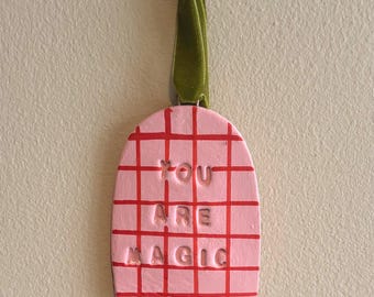 May include: A pink, grid-patterned ornament with the words "YOU ARE MAGIC" in raised letters. The ornament has a green ribbon for hanging. The grid lines and text are red.