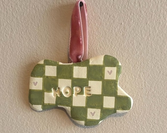 May include: A ceramic ornament with a green and white checkered pattern and the word "HOPE" in the center. The ornament has a pink ribbon for hanging and small heart details. The ornament is a decorative item.