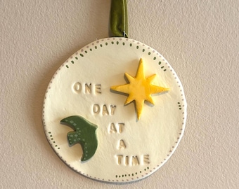 May include: A round, off-white ceramic wall hanging with the phrase "ONE DAY AT A TIME" in embossed lettering. It features a yellow star and a green bird, with a green ribbon for hanging. The border is dotted.