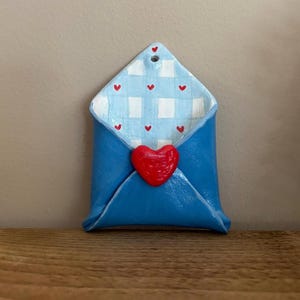 May include: A blue ceramic envelope with a red heart closure. The interior features a light blue and white gingham pattern with small red hearts. The envelope is designed with a hole at the top for hanging.