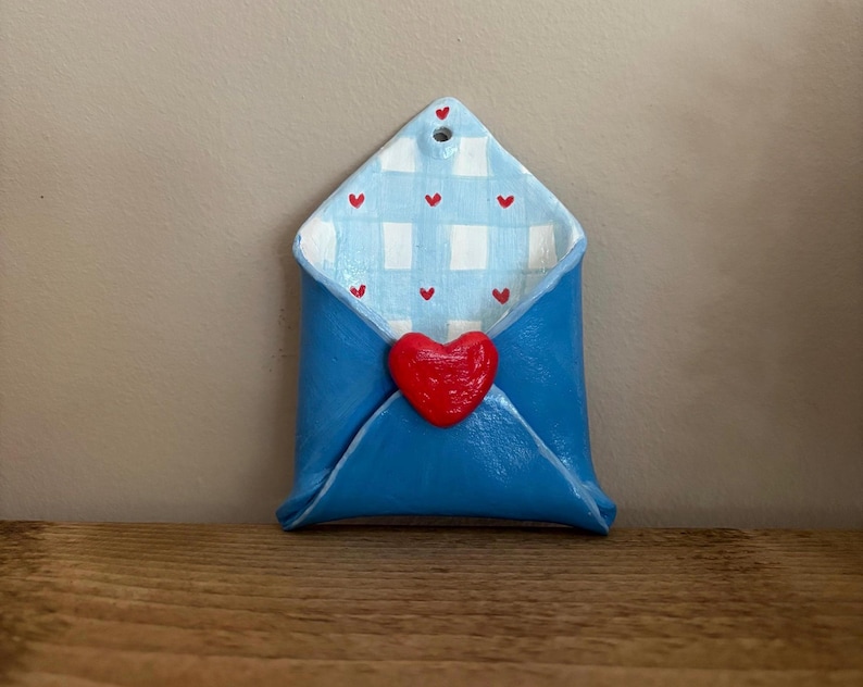 May include: A blue ceramic envelope with a red heart closure. The interior features a light blue and white gingham pattern with small red hearts. The envelope is designed with a hole at the top for hanging.