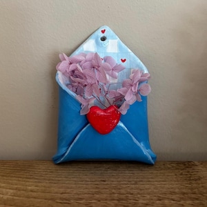 May include: A blue ceramic envelope-shaped wall hanging with a red heart and pink flowers. The envelope has a checkered pattern on the flap and small red heart accents. The flowers spill out of the envelope.