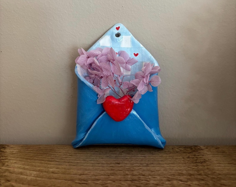 May include: A blue ceramic envelope-shaped wall hanging with a red heart and pink flowers. The envelope has a checkered pattern on the flap and small red heart accents. The flowers spill out of the envelope.