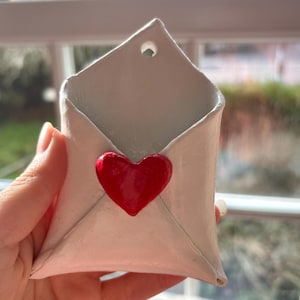 May include: A white ceramic envelope-shaped wall pocket with a bright red heart attached. The pocket has a hole at the top for hanging. The open design suggests it's for small items or decorative use. The background is a window.