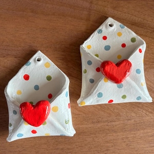 May include: Two white ceramic envelope-shaped wall pockets with red heart accents. The envelopes are decorated with colorful polka dots. Each has a small hole at the top for hanging. The pockets are displayed on a wooden surface.