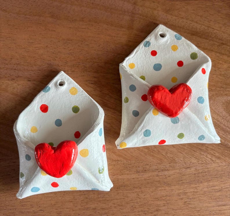 May include: Two white ceramic envelope-shaped wall pockets with red heart accents. The envelopes are decorated with colorful polka dots. Each has a small hole at the top for hanging. The pockets are displayed on a wooden surface.