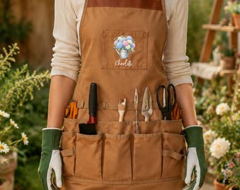 May include: A brown canvas apron with leather straps, featuring a chest pocket with a graphic of a watering can filled with flowers and the name Charlotte printed below. The apron includes multiple utility pockets holding various gardening tools.