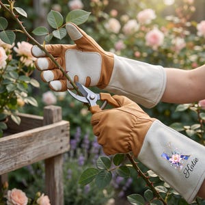 May include: Tan and cream leather gardening gloves with long gauntlet cuffs. The right cuff features a decorative letter K with a floral design and the name Katie printed in black script. The gloves are holding metal pruning shears.