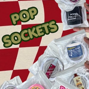 Peut inclure: Une collection de supports de téléphone circulaires sous emballage plastique transparent, présentant des répliques miniatures de divers produits dont le gel Ampro Pro Styl, le soin capillaire Blue Magic, le chewing-gum Hubba Bubba et le beurre de cacao Queen Helene.