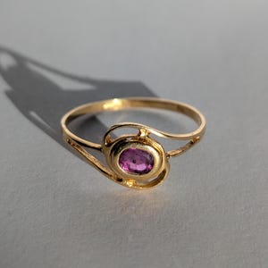 Può includere: Un anello d'oro con una gemma rosa ovale. L'anello presenta un design delicato e a spirale attorno alla gemma e una fascia sottile. L'anello è posto su uno sfondo grigio.