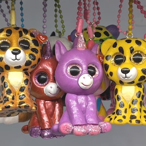 May include: A selection of colourful, glittery animal-themed plush toys, including a leopard, a unicorn, and a red creature. The leopard has a yellow body with black spots. The unicorn is purple with a pink horn. Each toy has large, glittery eyes.