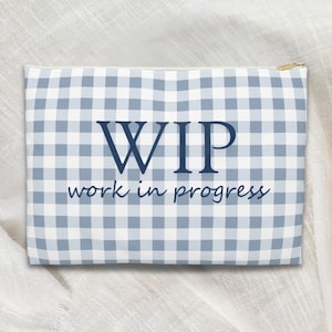 May include: A rectangular zippered pouch with a blue and white gingham pattern. The words "WIP work in progress" are printed in navy blue on the front. The zipper is gold.