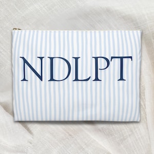 May include: A rectangular pouch with a light blue and white striped pattern. The pouch features the navy blue letters "NDLPT" across the front and a gold zipper. The pouch is likely made of fabric.