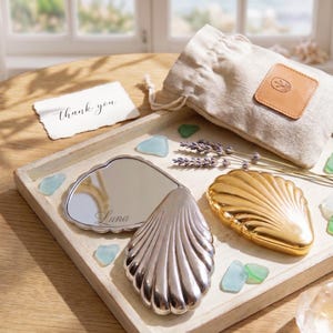May include: A decorative tray with a silver shell-shaped mirror engraved with "Luna", a gold shell-shaped compact, a small linen bag, and a card that says "Thank you". The tray is decorated with lavender sprigs and sea glass.