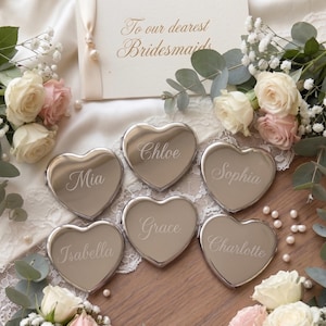 Könnte beinhalten: Sechs herzförmige, silberne Kompaktspiegel mit eingravierten Namen: Chloe, Mia, Sophia, Grace, Isabella und Charlotte. Eine Karte mit der Aufschrift "To our dearest Bridesmaids". Rosen und Grün umgeben die Spiegel auf einer Spitzentischdecke.