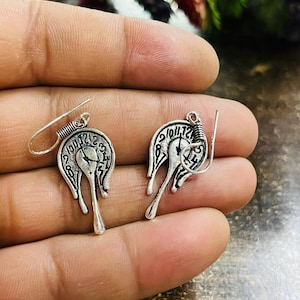 May include: A pair of silver-coloured earrings. Each earring has a clock face design with a melting effect, a wire-wrapped top, and a long, teardrop-shaped dangle. The earrings are shown against a neutral background.