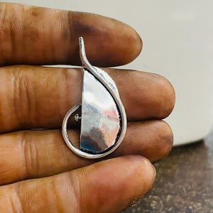 Op de afbeelding: Een zilveren hanger met een uniek ontwerp. De hanger heeft een halve maanvorm met een gebogen, bladachtig element en een klein, rond accent. Het metaal heeft een gepolijst, reflecterend oppervlak.