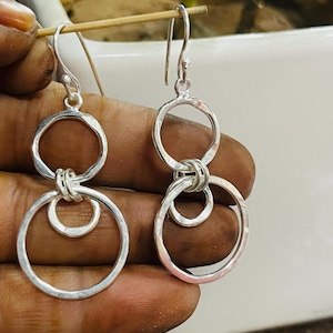 Puede incluir: Un par de pendientes colgantes de plata. Cada pendiente presenta dos círculos abiertos conectados, con un círculo más pequeño adjunto en la parte inferior. Los pendientes se suspenden mediante un gancho. El metal tiene una textura martillada.