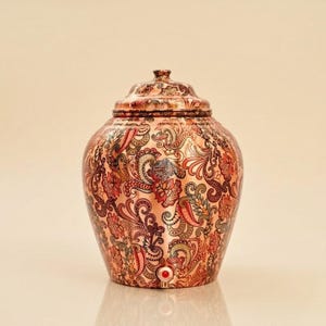 May include: A copper water dispenser with a rounded body and a matching lid. The surface features an intricate, multi-colored floral and paisley pattern in shades of red, green, and black. A small circular red accent is visible near the base.