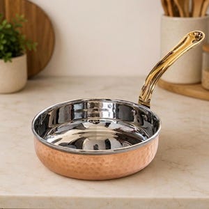 Könnte beinhalten: Eine runde Bratpfanne mit gehämmertem Kupferaußenbereich und poliertem, silberfarbenem Innenbereich. Die Pfanne hat einen langen, geschwungenen, reflektierenden goldfarbenen Metallgriff, der mit sichtbaren Nieten an der Seite befestigt ist.