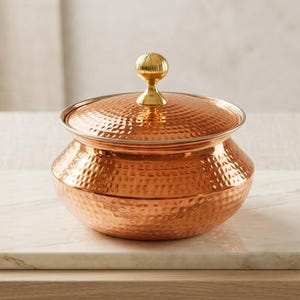 May include: A copper-colored, hammered metal serving bowl with a lid. The bowl has a rounded shape and a gold-colored knob on the lid. It sits on a white marble surface.