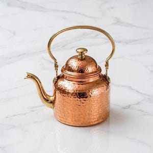 Puede incluir: Una tetera de cobre martillado con cuerpo redondeado, pico curvo y un asa de metal dorado pulido que se arquea sobre la parte superior. La tapa es abovedada con un pequeño pomo dorado a juego en el centro, ofreciendo un acabado metálico.