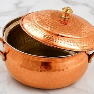 May include: A round, hammered copper pot with two brass side handles and a matching hammered copper lid featuring a small brass knob. The interior of the pot has a metallic finish, and the exterior displays a textured, dimpled surface.