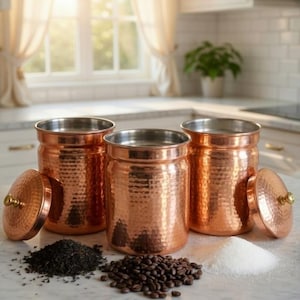 May include: Three copper-colored canisters with hammered texture and stainless steel interiors. Each canister has a matching lid with a gold-colored knob. Tea leaves, coffee beans, and sugar are arranged in front of the canisters. Kitchen storage.